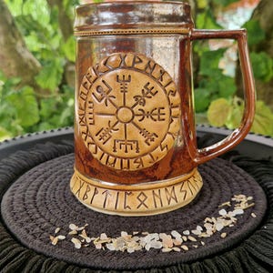 Op de afbeelding: Een bruine keramische mok met een cirkelvormig, in reliëf aangebracht Vegvisir-symbool omringd door runeninscripties. De mok heeft een glanzende afwerking, een stevig handvat en een lichtere basisband die ook is versierd met runentekens.