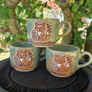 May include: Three ceramic mugs with a sage green glaze, each featuring a raised, hand-painted relief of a fox sitting next to a mushroom and a leafy branch. The mugs have a rounded shape, a sturdy handle, and a smooth, glossy finish throughout.