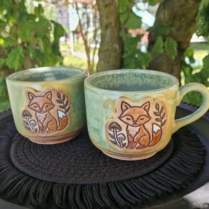 Könnte beinhalten: Zwei Keramikbecher mit hellgrüner und gelber Glasur. Jeder Becher zeigt ein erhabenes, geprägtes Design eines Fuchses, der neben kleinen Pilzen sitzt. Die Becher haben eine abgerundete Form, ein glattes Inneres und einen Henkel an der Seite.