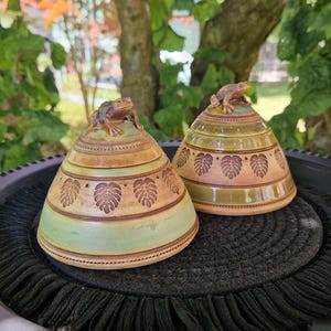 Könnte beinhalten: Zwei konische Keramikdosen mit Deckel in hellgrüner und beiger Glasur. Jeder Deckel ist mit einer modellierten Froschfigur verziert. Die Seiten der Dosen zeigen ein wiederkehrendes, geprägtes Blattmuster und horizontale braune Linien.