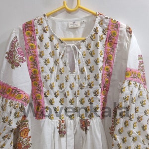 May include: White tunic dress with a floral print in yellow, pink, and green. The dress features a round neckline with a tie, long sleeves, and a pink border. The brand name "Wovenkala" is visible.