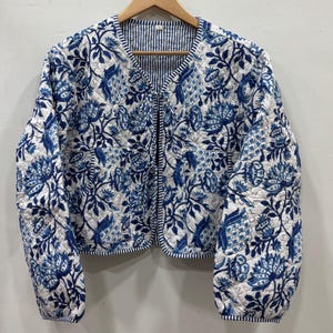 Puede incluir: Chaqueta acolchada con estampado floral azul y blanco. La chaqueta tiene mangas largas y cuello redondo. El forro interior presenta un patrón de rayas azules y blancas. La chaqueta está colgada en una percha de madera.