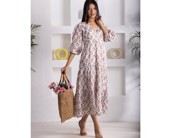 May include: A white dress with a floral print and a v-neckline. The dress has a tiered skirt and 3/4 sleeves. The model is holding a straw bag with a wooden handle.