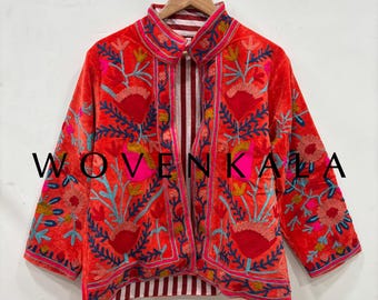 May include: A red embroidered jacket with floral patterns in pink, blue, and yellow. The jacket has a striped lining and is hanging on a wooden hanger. The text "WOVEN KALA" is visible on the front.
