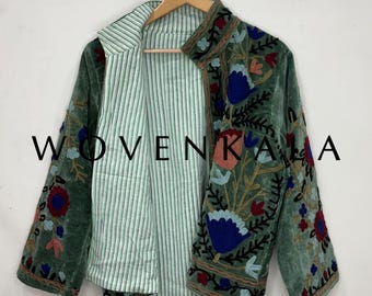May include: A green velvet jacket with floral embroidery in red, blue, and peach. The jacket has a striped lining and is hanging on a wooden hanger. The text "WOVENKALA" is visible on the left side of the jacket.