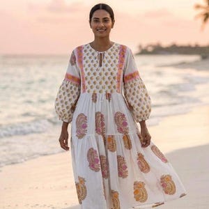 May include: A white midi dress with a paisley pattern in shades of pink and orange. The dress features long, puffy sleeves and a tie-neck detail. The model is walking on a sandy beach.