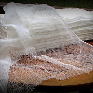May include: A pile of white gauze pads with a length of gauze draped across them. The gauze has a loose weave and a slightly frayed edge. The pads are neatly stacked on a wooden surface.