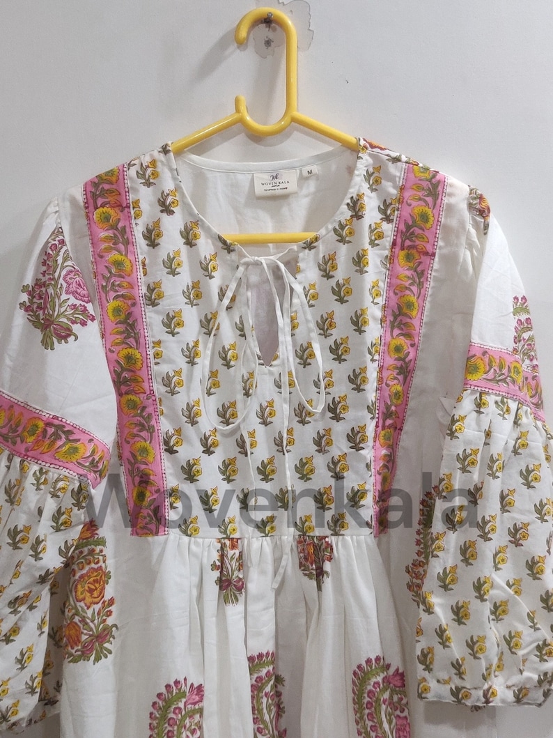 May include: White tunic dress with a floral print in yellow, pink, and green. The dress features a round neckline with a tie, long sleeves, and a pink border. The brand name "Wovenkala" is visible.