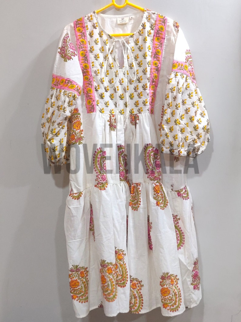 May include: White dress with a floral pattern in yellow, orange, and pink. The dress has long, puffy sleeves and a tiered skirt. The neckline features a tie closure. The text "WOVENKALA" is visible at the bottom of the dress.