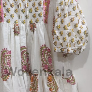May include: White dress with floral patterns in yellow, pink, and green. The dress features long, puffy sleeves and a tiered skirt. The neckline and sleeves are trimmed with pink fabric. The dress has a tie at the neckline.