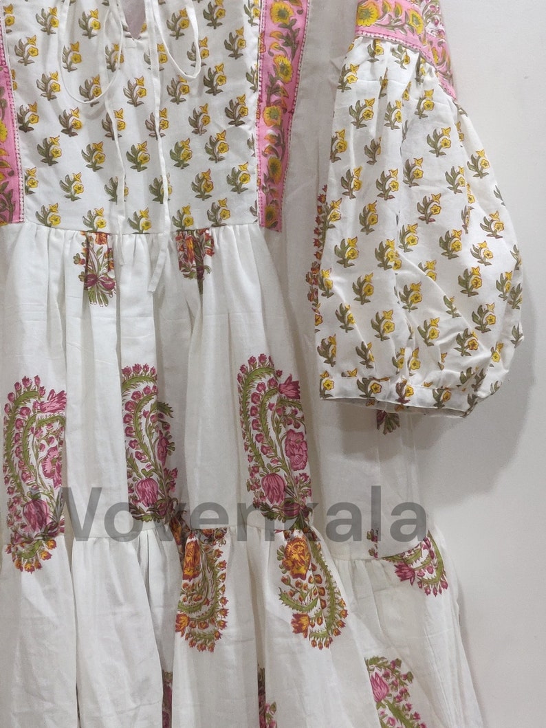 May include: White dress with floral patterns in yellow, pink, and green. The dress features long, puffy sleeves and a tiered skirt. The neckline and sleeves are trimmed with pink fabric. The dress has a tie at the neckline.
