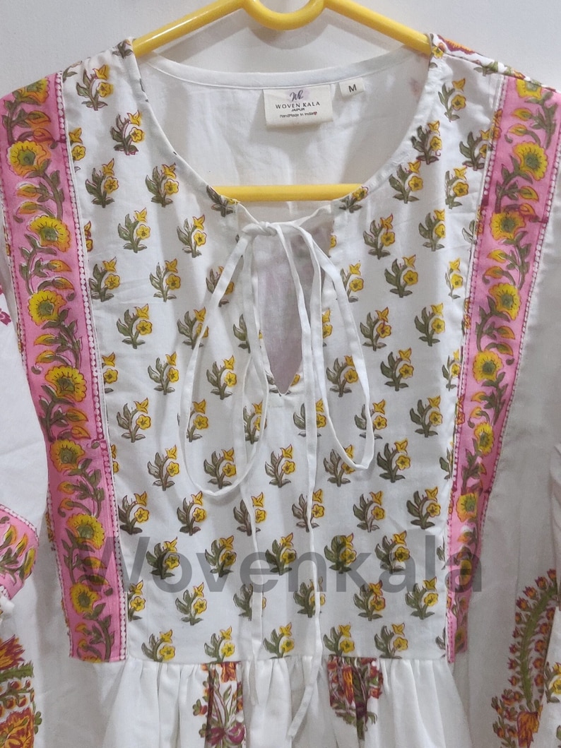 May include: White tunic with a floral print in yellow and green. The tunic has pink and yellow floral borders and a tie-up neckline. The brand "Woven Kala" is visible on a tag. The tunic is a size M.