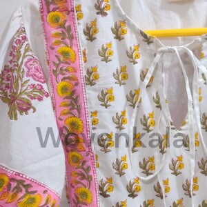 May include: White tunic top with a floral print. The top features a pink border with yellow flowers and green leaves. The sleeves have a floral pattern. The neckline has a tie closure. The tag reads "WOVEN KALA Jaipur Handmade in India."
