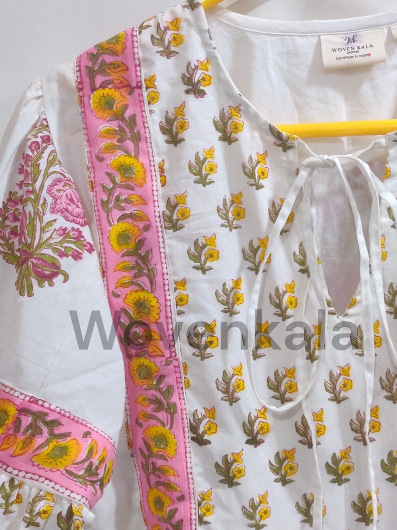 May include: White tunic top with a floral print. The top features a pink border with yellow flowers and green leaves. The sleeves have a floral pattern. The neckline has a tie closure. The tag reads "WOVEN KALA Jaipur Handmade in India."