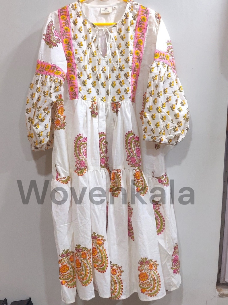 May include: White cotton dress with a floral print in yellow, pink, and orange. The dress features long, puffy sleeves and a tiered skirt. The neckline has a tie closure. The dress is a midi length.