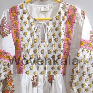 May include: White tunic with a floral pattern in yellow, pink, and green. The tunic has a tie-neck detail and long sleeves. The floral design is repeated on the sleeves and bodice.