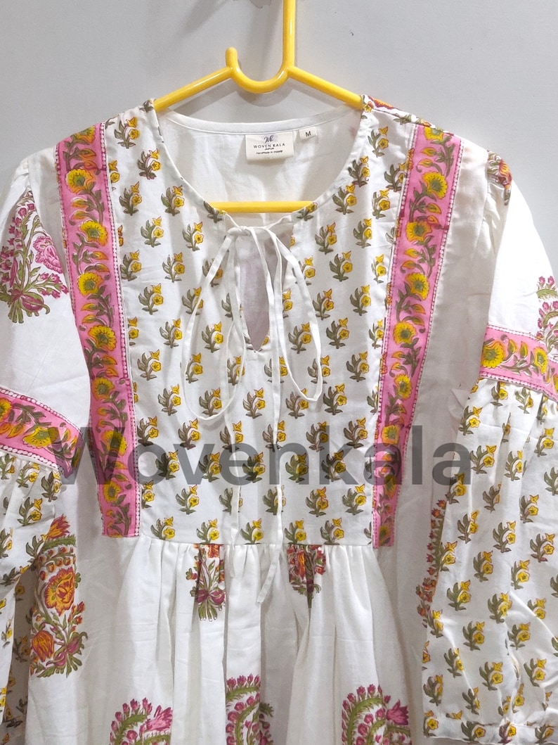 May include: White tunic with a floral pattern in yellow, pink, and green. The tunic has a tie-neck detail and long sleeves. The floral design is repeated on the sleeves and bodice.
