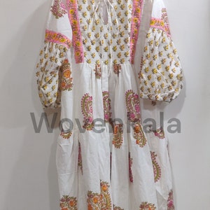 May include: White dress with floral patterns in yellow, orange, and pink. The dress features long, puffy sleeves and a tiered skirt. Pink and yellow trim accents the neckline and sleeves. The dress is hanging on a yellow hanger.