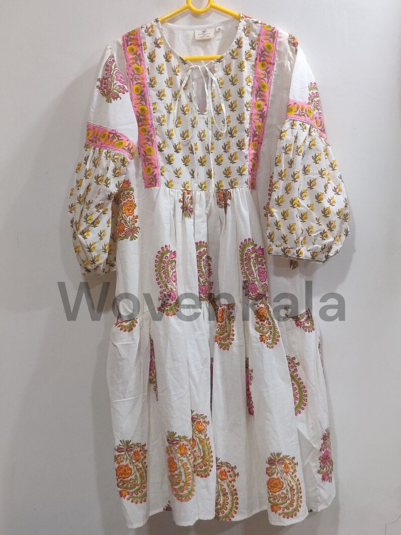 May include: White dress with floral patterns in yellow, orange, and pink. The dress features long, puffy sleeves and a tiered skirt. Pink and yellow trim accents the neckline and sleeves. The dress is hanging on a yellow hanger.