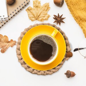 Puede incluir: Vista cenital de una taza y platillo amarillos con caf&eacute; oscuro, sobre un posavasos tejido. Hojas de oto&ntilde;o, bellotas, an&iacute;s estrellado, una bufanda amarilla y gafas evocan un ambiente oto&ntilde;al acogedor.