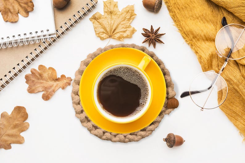 Puede incluir: Vista cenital de una taza y platillo amarillos con caf&eacute; oscuro, sobre un posavasos tejido. Hojas de oto&ntilde;o, bellotas, an&iacute;s estrellado, una bufanda amarilla y gafas evocan un ambiente oto&ntilde;al acogedor.