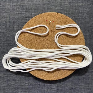 Puede incluir: Un tablero de corcho redondo con cuerda blanca y dos alfileres. La cuerda est&aacute; en bucle y enrollada, lo que sugiere un proyecto de artesan&iacute;a o bricolaje. El tablero de corcho es marr&oacute;n y la cuerda es blanca.