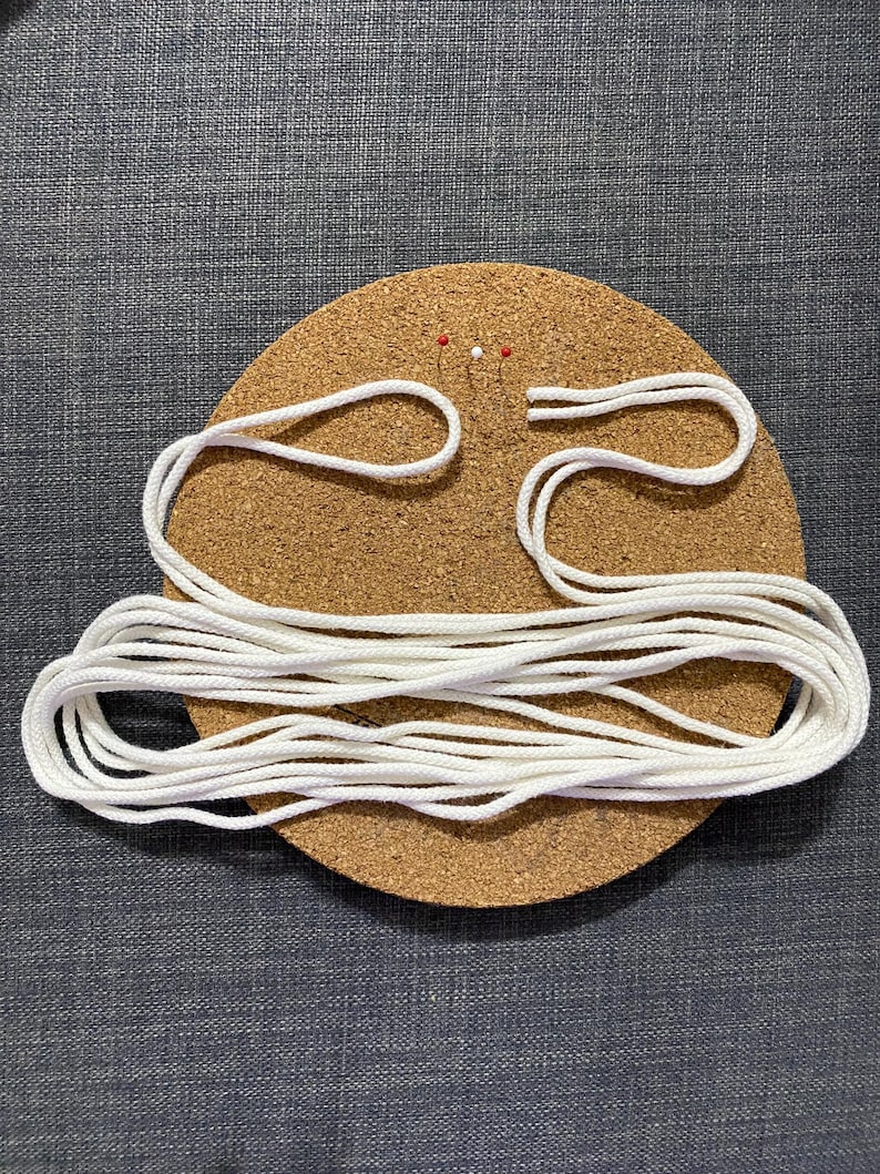 Puede incluir: Un tablero de corcho redondo con cuerda blanca y dos alfileres. La cuerda est&aacute; en bucle y enrollada, lo que sugiere un proyecto de artesan&iacute;a o bricolaje. El tablero de corcho es marr&oacute;n y la cuerda es blanca.