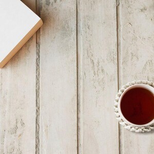 Puede incluir: Una taza de t&eacute; con asa rosa claro se encuentra sobre un posavasos de ganchillo blanco. Un libro cerrado con cubierta blanca y bordes beige est&aacute; en la esquina superior izquierda. Los objetos est&aacute;n sobre una superficie de madera clara.