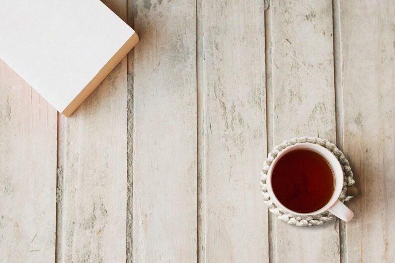 Puede incluir: Una taza de t&eacute; con asa rosa claro se encuentra sobre un posavasos de ganchillo blanco. Un libro cerrado con cubierta blanca y bordes beige est&aacute; en la esquina superior izquierda. Los objetos est&aacute;n sobre una superficie de madera clara.