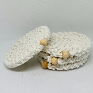 以下が含まれることがあります： オフホワイトのかぎ針編みコースター、木製ビーズのアクセント付き。コースターは丸く積み重ねられており、1つがスタックに寄りかかっています。かぎ針編みは質感のある外観です。木製ビーズはライトブラウンです。