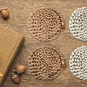 Puede incluir: Cuatro posavasos circulares hechos a mano, en tonos beige y blanquecino, con un aspecto texturizado y tejido. Los posavasos est&aacute;n dispuestos sobre una superficie de madera, junto con hojas de roble secas, bellotas y un libro cerrado.