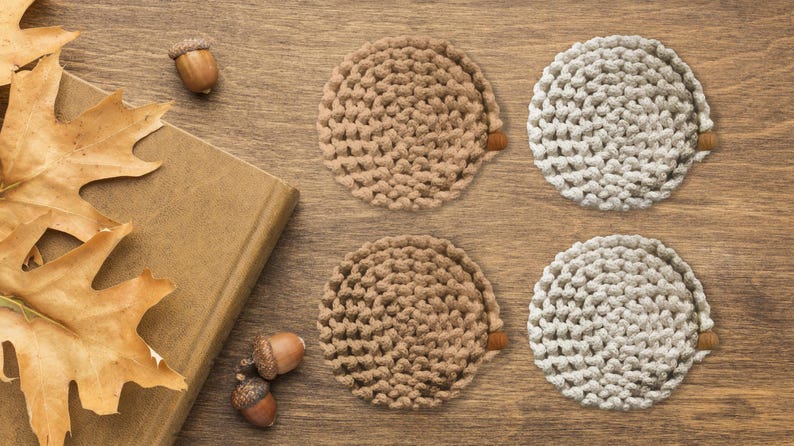Puede incluir: Cuatro posavasos circulares hechos a mano, en tonos beige y blanquecino, con un aspecto texturizado y tejido. Los posavasos est&aacute;n dispuestos sobre una superficie de madera, junto con hojas de roble secas, bellotas y un libro cerrado.
