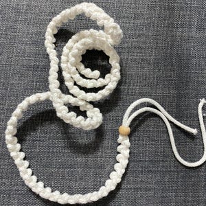 Puede incluir: Un cord&oacute;n de macram&eacute; blanco con una cuenta de madera, dispuesto en forma circular, descansa sobre una superficie gris texturizada. El cord&oacute;n est&aacute; anudado y en bucle, con una porci&oacute;n enrollada y otra formando un lazo.