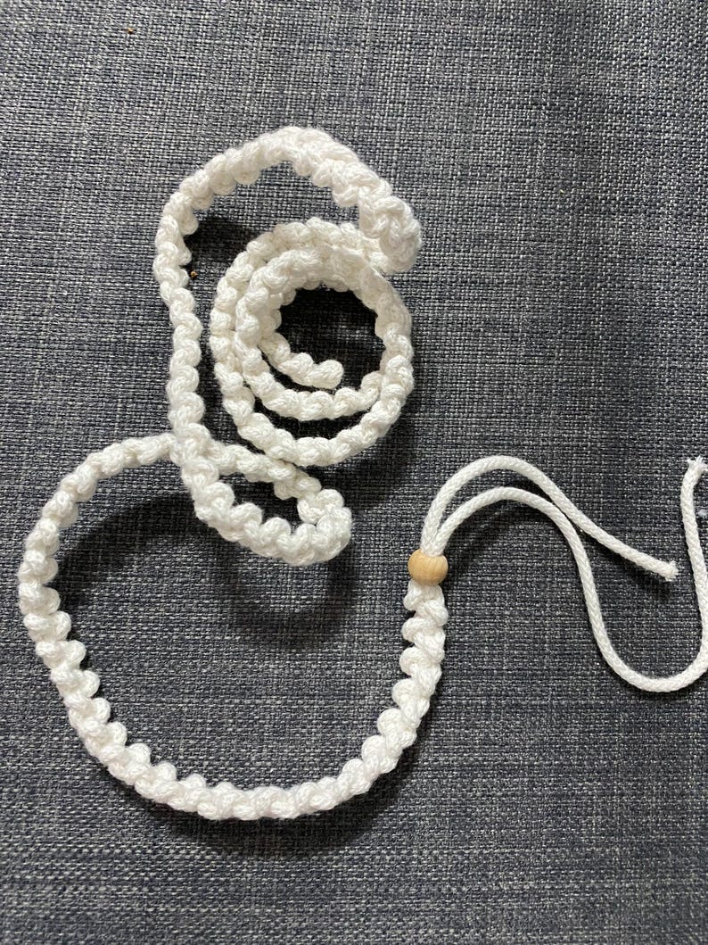 Puede incluir: Un cord&oacute;n de macram&eacute; blanco con una cuenta de madera, dispuesto en forma circular, descansa sobre una superficie gris texturizada. El cord&oacute;n est&aacute; anudado y en bucle, con una porci&oacute;n enrollada y otra formando un lazo.