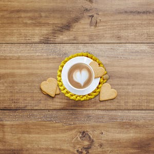 Puede incluir: Una taza de caf&eacute; blanca con un dise&ntilde;o en forma de coraz&oacute;n en la espuma se encuentra sobre un posavasos amarillo. Galletas en forma de coraz&oacute;n est&aacute;n dispuestas alrededor de la taza sobre una superficie de madera. La escena evoca un ambiente acogedor y rom&aacute;ntico.