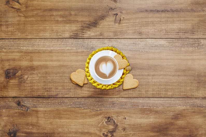 Puede incluir: Una taza de caf&eacute; blanca con un dise&ntilde;o en forma de coraz&oacute;n en la espuma se encuentra sobre un posavasos amarillo. Galletas en forma de coraz&oacute;n est&aacute;n dispuestas alrededor de la taza sobre una superficie de madera. La escena evoca un ambiente acogedor y rom&aacute;ntico.