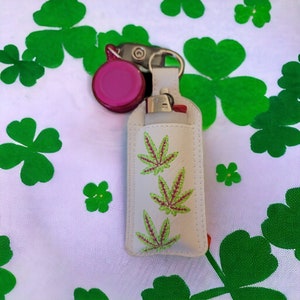 May include: White leather lighter case with a green and pink marijuana leaf design. The case has a pink keychain attached.