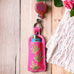 May include: Pink leather lighter case with a green marijuana leaf design and a retractable keychain. The case is attached to a silver keychain with a pink retractable reel.