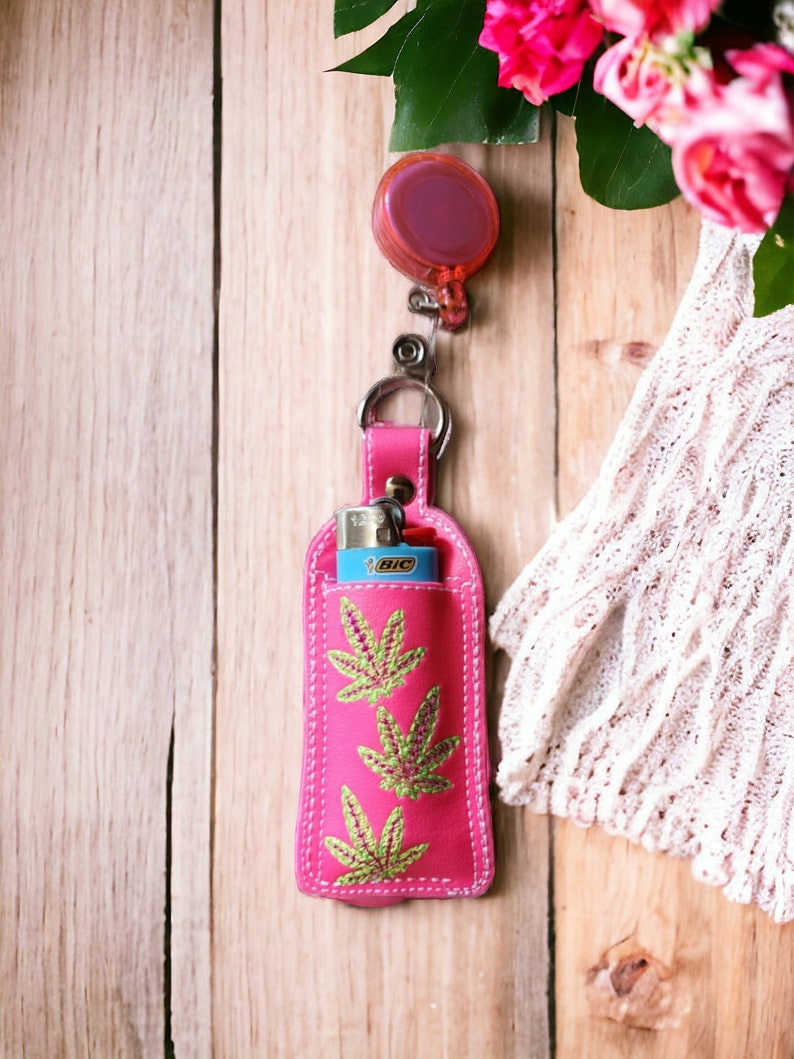 May include: Pink leather lighter case with a green marijuana leaf design and a retractable keychain. The case is attached to a silver keychain with a pink retractable reel.