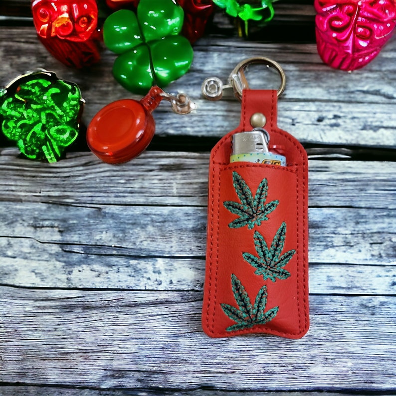 May include: A red leather lighter case with a zipper closure and a key ring. The case is decorated with three green embroidered marijuana leaf designs.