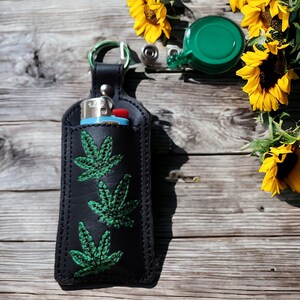 May include: Black leather keychain with a green embroidered marijuana leaf design. The keychain has a metal ring and a small blue lighter inside.