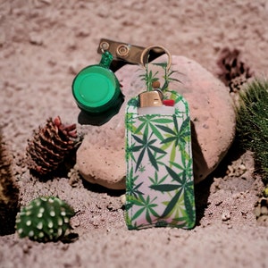 May include: A green and white patterned keychain with a small, clear container and a lighter inside. The keychain has a leaf design.