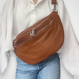 May include: A brown leather fanny pack with silver zippers and a silver chain strap. The bag is worn around the waist of a person wearing a white shirt and blue jeans.