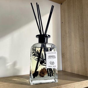 May include: A clear glass diffuser bottle with black reeds and a black cap. The bottle contains botanical elements and the text "Jasmine & Little Paleo" and "Cul Tural". The bottle is 200 ml / 6.8 fl oz.