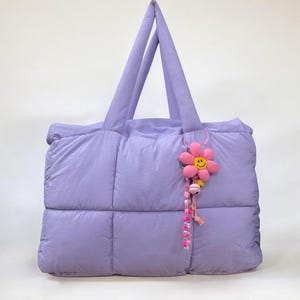 May include: A lavender quilted tote bag with a pink flower charm with a smiley face. The charm is attached to a pink beaded chain.