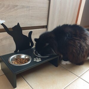May include: Black cat silhouette pet food stand with two stainless steel bowls. One bowl is filled with dry cat food. A black and white cat is eating from the other bowl.