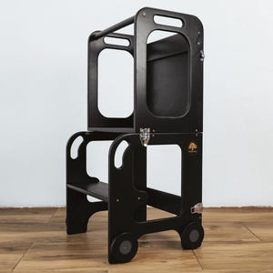 May include: Black wooden learning tower with two steps and a safety rail. The tower has wheels and a small shelf on the top.