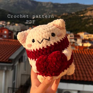 May include: Crochet pattern PDF for a white and red amigurumi plush toy holding a red heart. The toy has a cute cartoon face with black eyes and a pink nose.