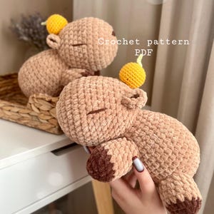 Puede incluir: Dos juguetes de peluche marrones tejidos a crochet con fruta amarilla tejida a crochet en la parte superior. Los juguetes tienen la forma de un capibara. El texto "Crochet pattern PDF" es visible en la imagen.