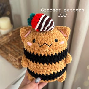 May include: Crochet pattern PDF for a brown and black striped amigurumi toy with a red and white strawberry on top.